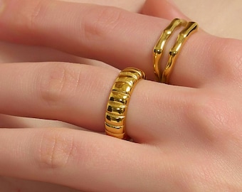 Könnte beinhalten: Zwei goldene Ringe werden auf einer Hand gezeigt. Ein Ring hat ein geripptes Design, während der andere ein doppelt gebogenes Design hat. Die Ringe bestehen aus glänzendem, gelbem Metall. Der Hintergrund ist eine verschwommene Ansicht von Haut und Kleidung.