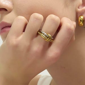 May include: A gold and silver ring with a knot design, featuring a band of small, clear stones. A gold hoop earring is visible in the ear. The ring is on a finger, and the hand is near the face.