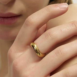 May include: A gold-colored ring with a smooth, curved design. The ring is worn on a finger, and the image shows a close-up of the hand and ring. The ring has a polished finish and reflects light. The jewelry is a simple, elegant accessory.