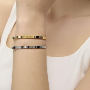 May include: A gold ring and hoop earrings are visible on a person's hand and ear. Two bracelets, one gold and one silver, are on the wrist. The bracelets and ring have a geometric design. The person is wearing a white vest top.
