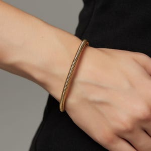 May include: A gold-coloured, textured bracelet is worn on a person's wrist. The bracelet has a ribbed design and a smooth, polished finish. The background is a neutral grey, and the person is wearing a black garment.