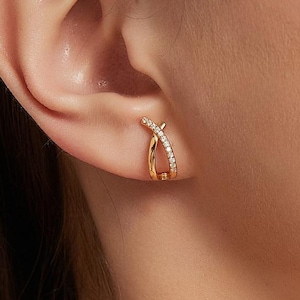 以下が含まれることがあります： 金色のフープピアス、クロスデザイン。 ピアスの片側には、小さな輝く石が飾られています。 ピアスは、色白の人の耳に着用されています。