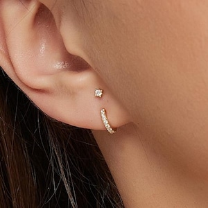 May include: Close-up of a person's ear adorned with two gold earrings. One is a small stud with a single clear stone, and the other is a small hoop earring with multiple clear stones. The earrings are set against the backdrop of the person's skin and hair.