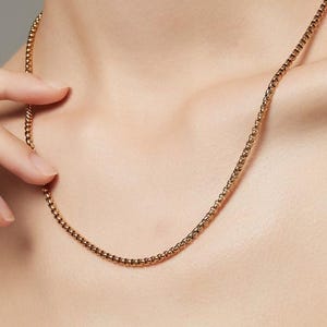 May include: A gold-coloured chain necklace with a delicate, beaded design. The necklace is worn around the neck and rests on the chest. The background is a neutral grey.