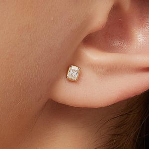 Könnte beinhalten: Ein goldfarbener Ohrstecker mit einem rechteckigen, klaren Edelstein. Der Ohrring ist in einer goldenen Lünette gefasst und wird am Ohr getragen. Das Bild ist eine Nahaufnahme des Ohrrings und des Ohrs.