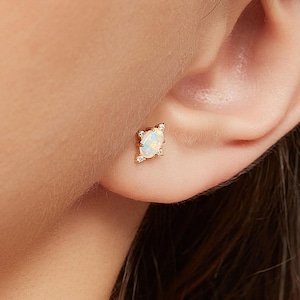Peut inclure: Boucle d'oreille en or avec une opale ovale au centre, entourée de quatre petites pierres claires. La boucle d'oreille est portée sur une oreille, mettant en valeur le design et les détails.