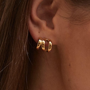 May include: Close-up of a pair of gold-colored hoop earrings. The earrings have a unique design, with a double-hoop appearance. The earrings are shiny and reflect light, suggesting a polished finish. The earrings are worn on an ear.