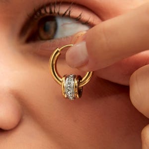 Pode incluir: Um brinco de argola dourado com um detalhe decorativo em prata e pedras transparentes. O brinco é segurado perto de um olho, mostrando o design e os detalhes da joia. O design inclui um aro dourado e uma faixa prateada com pedras transparentes.