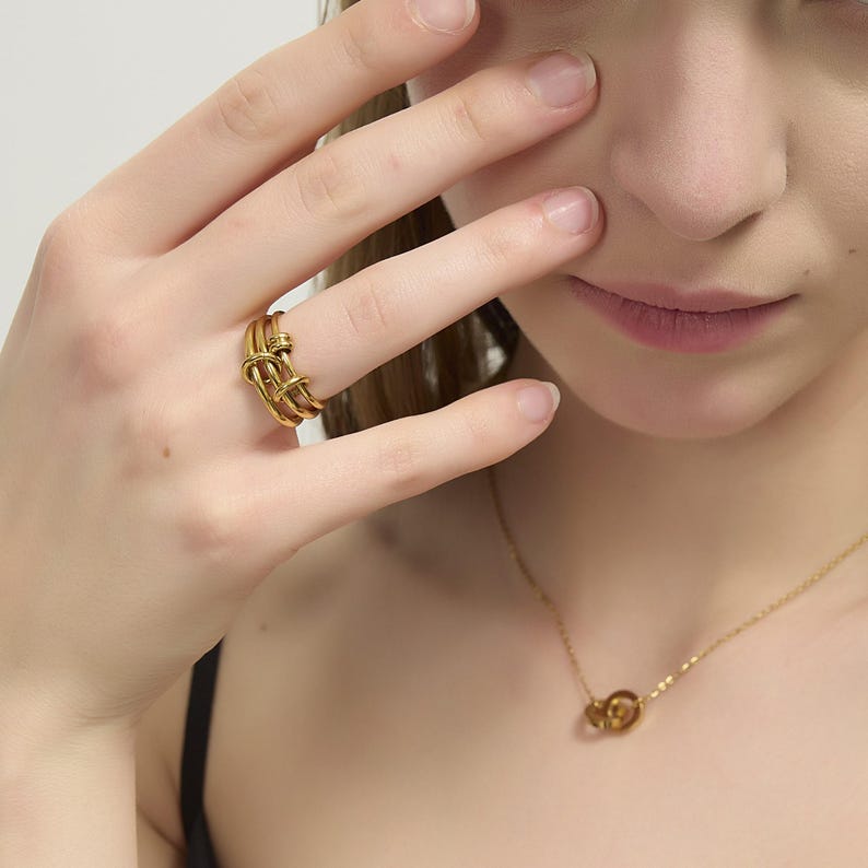 May include: A gold-toned ring with a knotted design. The ring is worn on the index finger of a person's hand.