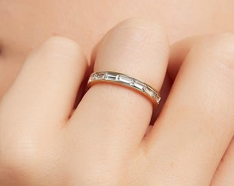 May include: A gold band ring set with rectangular-cut, clear gemstones. The ring is worn on a finger, showcasing the delicate design and sparkle of the gemstones. The ring's setting is a warm gold color, complementing the clear gemstones.