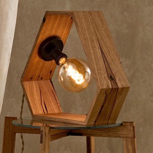 May include: A hexagonal wooden lamp with a light bulb inside. The lamp is made of reclaimed wood and has a rustic look. It is sitting on a wooden table with a glass top.