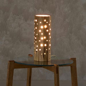 May include: A wooden table lamp with a cylindrical base and a pattern of small holes. The lamp is lit and casting a warm glow. The lamp is sitting on a glass-topped table with wooden legs.