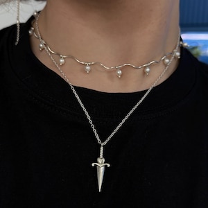 Könnte beinhalten: Silbernes Halsketten-Set mit einem filigranen Dolch-Anhänger mit herzförmigem Griff. Das Set beinhaltet ein wellenförmiges Choker-Halsband mit kleinen Perlen. Der Schmuck wird vor schwarzem Hintergrund präsentiert.