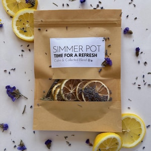 May include: A brown paper bag labeled "Simmer Pot" with the text "Time for a Refresh" and "Calm & Collected Blend." The bag contains dried lemon slices, lavender, and rosemary. Sliced lemons and dried flowers are scattered around the bag.