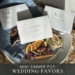 May include: Mini simmer pot wedding favors with dried orange slices, cinnamon sticks, and rosebuds. Each favor is packaged in a clear bag with a personalized card. Text on the cards includes names, dates, and "Thank You".
