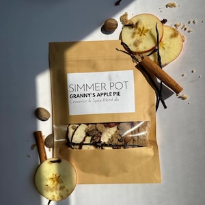May include: A tan paper pouch labelled SIMMER POT GRANNY'S APPLE PIE Cinnamon & Spice Blend with a clear window showing dried apple slices, cinnamon sticks, and brown sugar. The pouch is surrounded by fresh apple slices, whole nutmeg, and cloves.