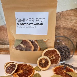 May include: A brown paper bag labeled "Simmer Pot Sunny Days Ahead Fruity & Fresh Blend" with dried orange slices, bay leaves, and a small glass bowl of lavender buds. The bag has a clear window showing the contents.