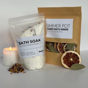 May include: A clear bag of bath soak with the text "BATH SOAK Oat Milk Mineral Soak" next to a brown paper bag with the text "SIMMER POT SUNNY DAYS AHEAD Fruity & Fresh Blend". A lit candle and dried orange slices are also visible.