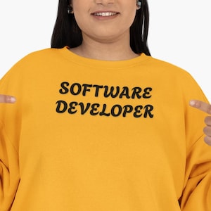 Könnte beinhalten: Eine Frau trägt ein gelbes Sweatshirt mit dem Text "SOFTWARE DEVELOPER" in Schwarz. Sie zeigt mit beiden Händen auf den Text auf ihrem Sweatshirt.
