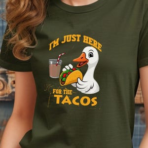 May include: Dark green t-shirt with a cartoon duck holding a taco and a drink. The text on the shirt reads "I'M JUST HERE FOR THE TACOS". The design is in white, orange, and yellow.