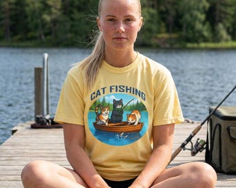 May include: A yellow t-shirt with the words "CAT FISHING" above an illustration of three cats in a boat fishing. The cats are orange, black, and orange and white. The person is sitting on a wooden dock.