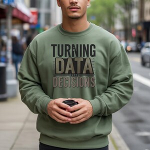 May include: An olive green sweatshirt featuring the text "TURNING DATA INTO DECISIONS". The text is in a distressed font. The person in the image is holding a black coffee cup. The sweatshirt is a crew neck style.