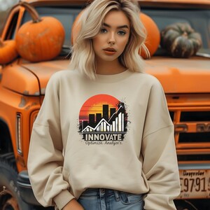 Peut inclure: Un sweat-shirt beige avec un motif graphique représentant un graphique ascendant dans un coucher de soleil. Le texte "INNOVATE" est affiché en dessous du graphique, avec "Optimize. Analyze's." en dessous. Le fond comprend des citrouilles et un camion orange.