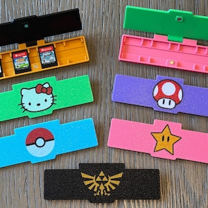 May include: A collection of colorful game cartridge holders. Designs include a Hello Kitty face, a Pokeball, a Super Mario mushroom, and a gold star. One holder is black with a gold Triforce symbol, and another holds three game cartridges.