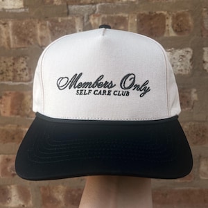 May include: A beige and black Members Only Self Care Club hat. The hat has a black brim and a beige crown with the words "Members Only" and "Self Care Club" embroidered in black script.