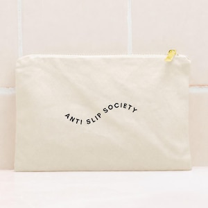 K&ouml;nnte beinhalten: Cremefarbene Canvas-Tasche mit goldenem Rei&szlig;verschluss und dem schwarzen Aufdruck "ANTI SLIP SOCIETY". Die rechteckige Tasche scheint aus einem weichen Stoff zu sein.