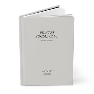 May include: A pale grey journal with the text "PILATES SOCIAL CLUB MEMBERS ONLY" printed in black. The journal also has the name "NICOLETTE" and a decorative flourish at the bottom. The pages are white.