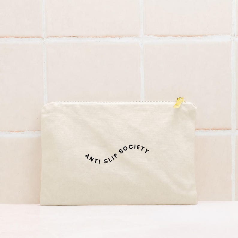K&ouml;nnte beinhalten: Cremefarbene Canvas-Tasche mit goldenem Rei&szlig;verschluss und dem schwarzen Aufdruck "ANTI SLIP SOCIETY". Die rechteckige Tasche scheint aus einem weichen Stoff zu sein.