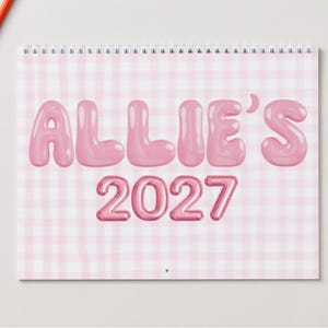 Puede incluir: Un calendario de pared con encuadernación en espiral y un fondo de patrón de cuadros vichy en rosa claro y blanco. El texto ALLIE'S 2027 está impreso en el centro con una fuente grande y redondeada con aspecto rosa brillante y tridimensional.