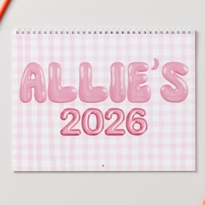 May include: A spiral-bound calendar with a pink gingham pattern background. The text "ALLIE'S 2026" is displayed in a bubble-style font. A red pencil and an orange accessory are visible in the background.