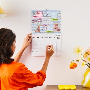 May include: A wall calendar for 2026 with a blue background and illustrations. The calendar has a spiral binding and is being written on with a pencil. The top of the calendar has the text "Make America Cowart Again" and "Girls Just Wanna Have Fun".