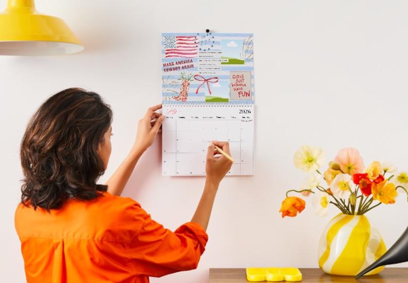 May include: A wall calendar for 2026 with a blue background and illustrations. The calendar has a spiral binding and is being written on with a pencil. The top of the calendar has the text "Make America Cowart Again" and "Girls Just Wanna Have Fun".