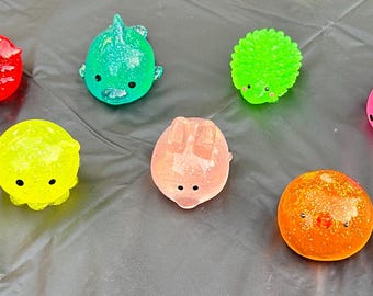 Op de afbeelding: Een verzameling kleurrijk, zacht speelgoed in verschillende vormen, waaronder een rode stekelbal, een groene dinosaurus en een roze vis. Het speelgoed is gerangschikt op een donkergrijze ondergrond.