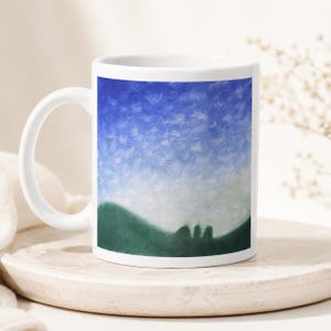May include: A white ceramic mug featuring a printed landscape illustration. The design displays a vibrant blue sky filled with textured white clouds above a soft, rolling green hill silhouette that spans the lower portion of the mug surface.