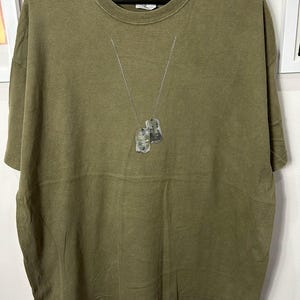 以下が含まれることがあります： クルーネックと半袖のオリーブグリーンのTシャツ。2つのドッグタグが付いたシルバーチェーンがネックラインからぶら下がっています。シャツは白い背景に黒いハンガーに吊るされています。