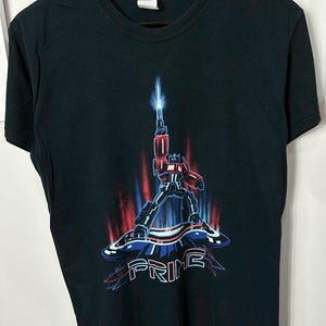 May include: Black t-shirt featuring a graphic of Optimus Prime, a red and blue robot, holding a weapon with a blue beam. The word "PRIME" is displayed in blue and white. The shirt is hanging on a black hanger.