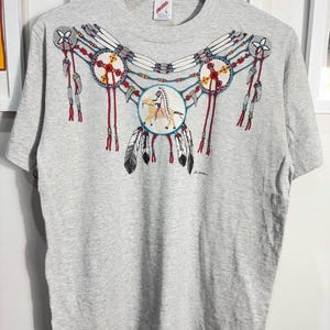 Peut inclure: Un t-shirt gris avec un motif d'inspiration amérindienne représentant un collier de perles avec des plumes et un cavalier à cheval. Le motif est dans des tons de rouge, bleu, blanc et noir.