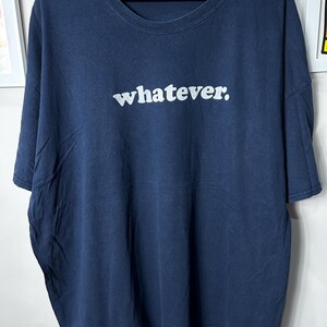 Op de afbeelding: Marineblauw T-shirt met het woord "whatever." in het wit over de borst gedrukt. Het shirt heeft korte mouwen en een ronde hals. Het shirt hangt aan een zwarte hanger.
