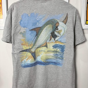 Op de afbeelding: Een grijs t-shirt met een levendige illustratie van een springende tarponvis. Het kunstwerk toont de vis in de lucht boven het water, met een achtergrond van een zonsondergang en wolken. Het shirt is gemaakt van een zacht materiaal.
