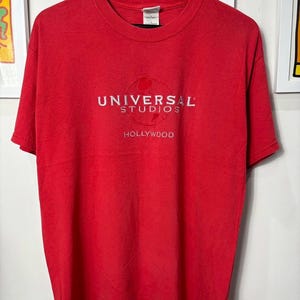 Op de afbeelding: Een rood t-shirt met het witte Universal Studios Hollywood-logo. Het logo bevat de woorden "UNIVERSAL STUDIOS" boven "HOLLYWOOD". Het shirt heeft korte mouwen en lijkt van een zacht materiaal te zijn.