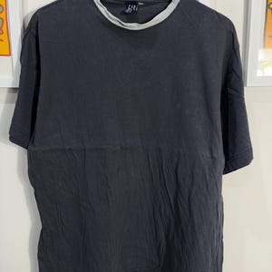 Peut inclure: Un t-shirt noir avec un col côtelé gris. Le t-shirt a une petite étiquette bleue avec le mot "GAP" dessus.