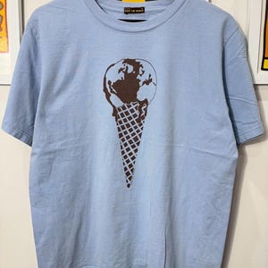 May include: Light blue t-shirt featuring a brown graphic of the Earth as an ice cream cone. The shirt has short sleeves and a crew neck. The text "SAVE THE WORLD" is printed on the collar.