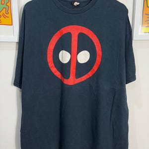 Könnte beinhalten: Ein dunkelblaues T-Shirt mit einem roten Deadpool-Logo auf der Vorderseite. Das Logo ist ein Kreis mit einem roten Schrägstrich und zwei weißen Augen.