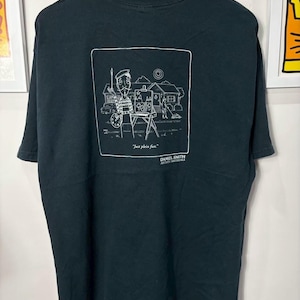 May include: Black t-shirt featuring a white illustration of an artist painting a landscape. The image includes the text "Just plein fun." and "DANIEL SMITH ARTIST MATERIALS". The shirt is hanging on a black hanger.