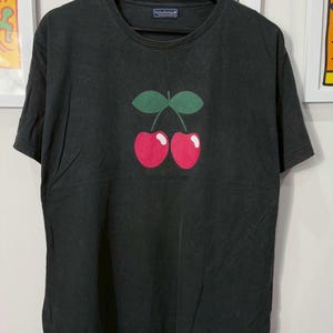 Puede incluir: Camiseta negra con un gráfico de dos cerezas rojas y hojas verdes. La camiseta tiene cuello redondo y mangas cortas. El nombre de la marca "Pacha Brothers" es visible en la etiqueta.