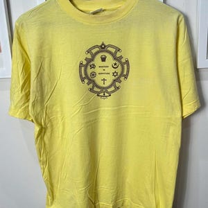 以下が含まれることがあります： クルーネックと半袖のイエローTシャツ。シャツには、「Mastery in Servitude」の文字とさまざまな宗教的シンボルが描かれた茶色の円形グラフィックが特徴です。シャツは柔らかく軽い生地で作られています。